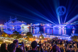 Das Bild zeigt ein Friedenssymbol, das während einer Show der Hamburg Cruise Days von Drohnen gebildet wurde sowie die aida prima und viele Besuchende im Hamburger Hafen.