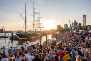 Das Bild zeigt den Großsegler Rickmer Rickmers im Hamburger Hafen sowie viele Besuchende der Hamburg Cruise Days entlang der Flutschutzmauer bei Sonnenuntergang.