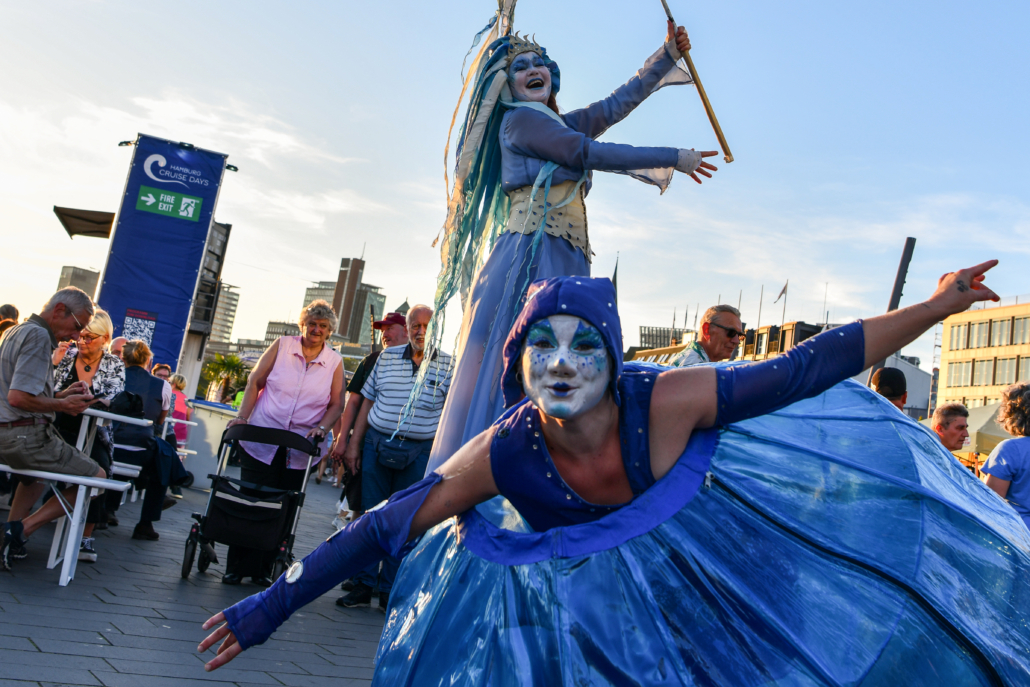 Das Bild zeigt zwei Menschen auf Stelzen, Besuchende und einen Informationsturm auf den Cruise Days 2023.