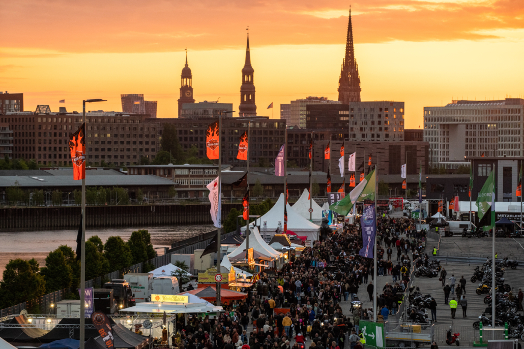 Das Bild zeigt die Veranstaltungsfläche der Hamburg Harley Days mit Ständen und Besuchenden vor einem schönen Sonnenuntergang.